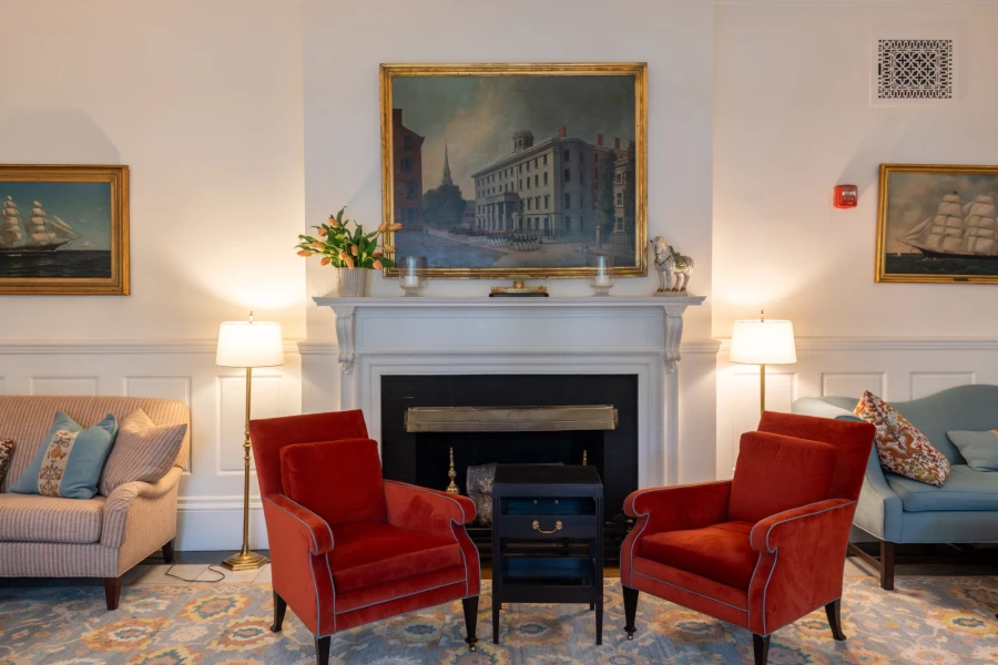Two red velvet chairs placed in front of a fireplace. Paintings in gold frames line the walls behind.